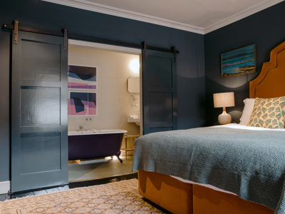 A bedroom with mustard coloured bed, dark grey walls and open door revealing a roll-top bath in the en-suite bathroom. 