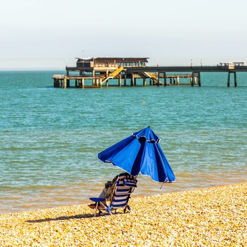 A blue and white striped deckchair with blue parasol, on a shingle beach with the sea and pier