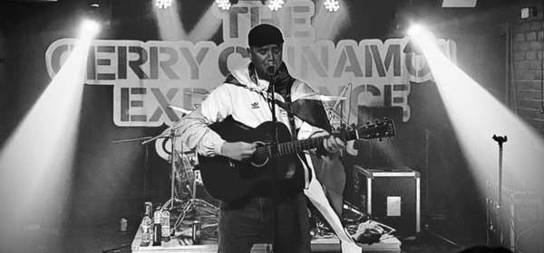 Photo of Gerry Cinna-Man performing on stage singing and playing a guitar.