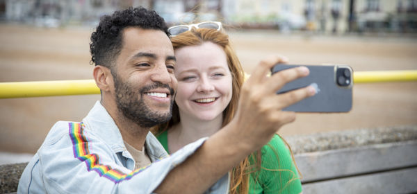 Two people taking a selfie with a smartphone with Deal beach and seafront in the background.