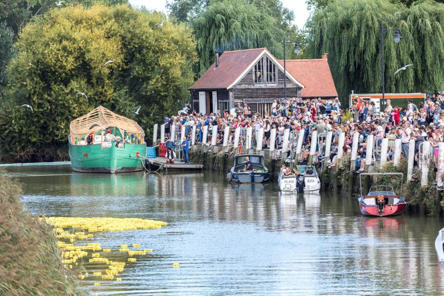 A crowd of people on a riverbank, yellow plastic ducks in the river