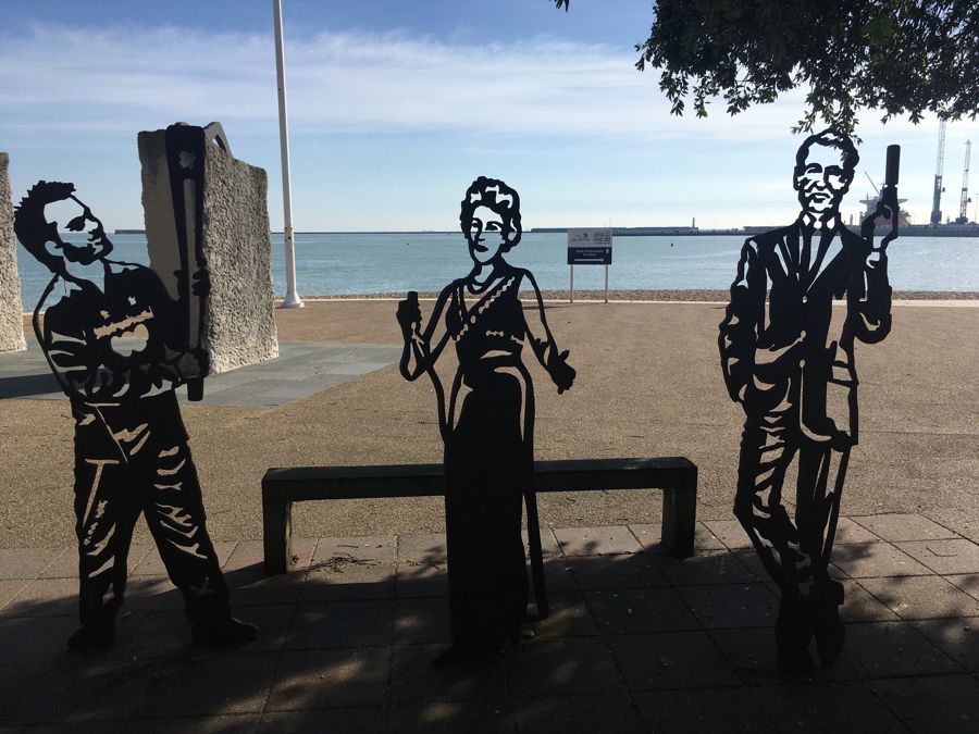 Three black metallic statues - one of Ian Fleming - with Dover Harbour behind.