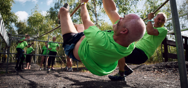 A group wearing green t-shirts navigate an outdoor obstacle course. The man in the foreground is hanging upside down from a rope.